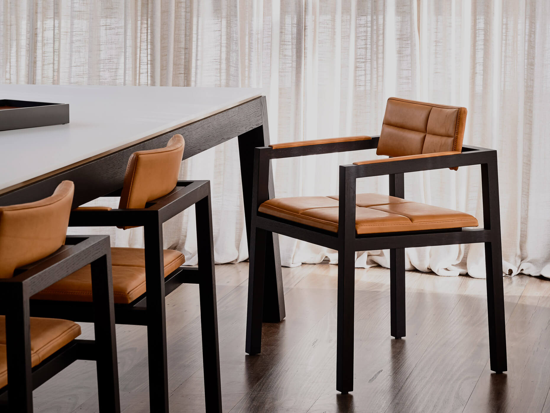 Soft linen curtains dapple light over beautiful designer dining table and chair set in soft tan leather